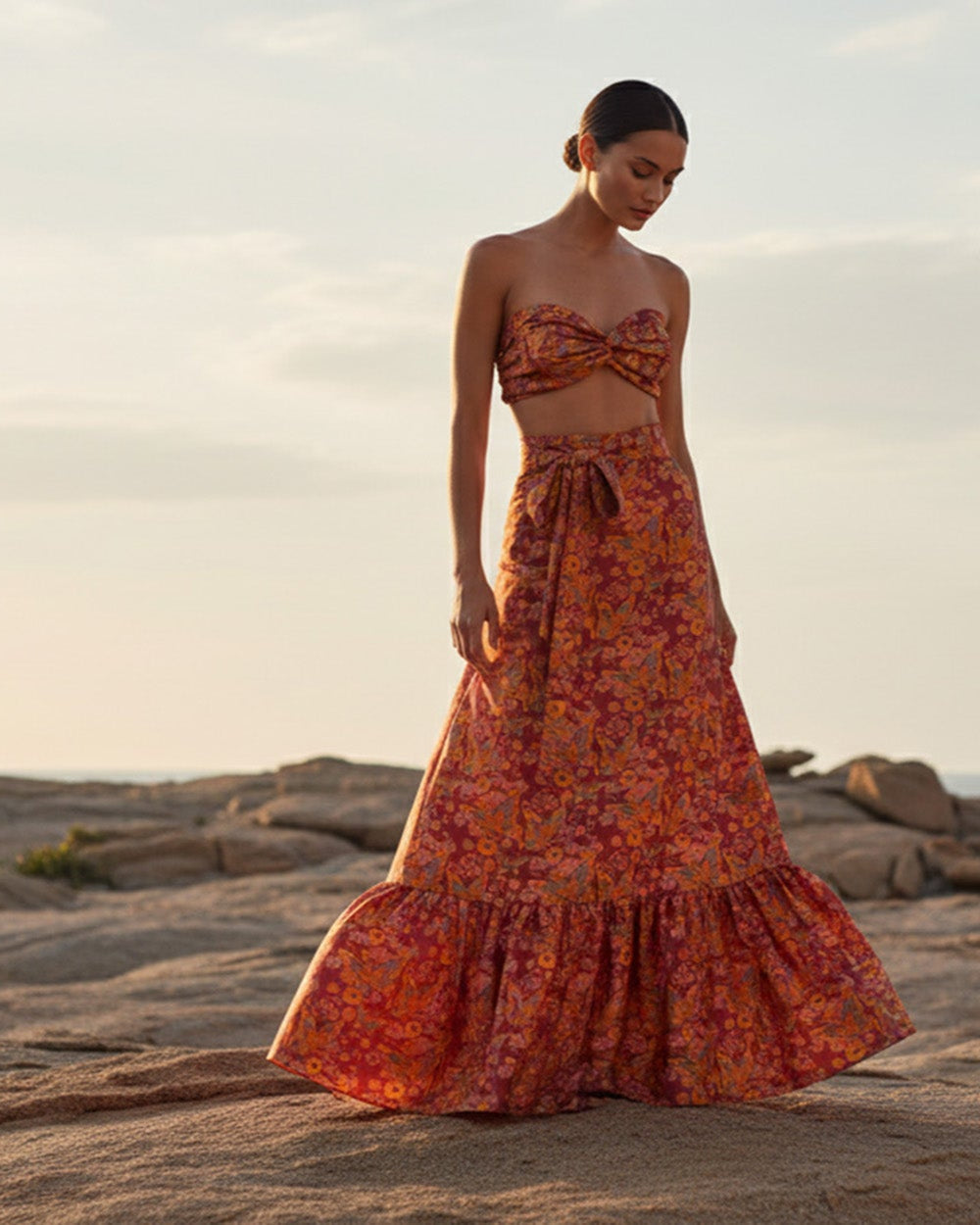 Bia Bandeau & Calypso Skirt in Silk. Woman in a floral dress standing on a rocky beach at sunset. Bandeau & Calypso Skirt